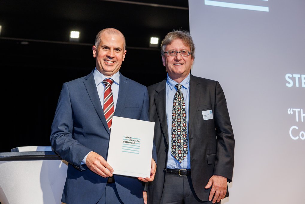 Stephen Casale and Jan-Michael Rost in suits stand side by side, Stephen holding a folder labeled "Max Planck Schools."