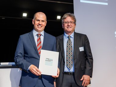 Stephen Casale and Jan-Michael Rost in suits stand side by side, Stephen holding a folder labeled "Max Planck Schools."