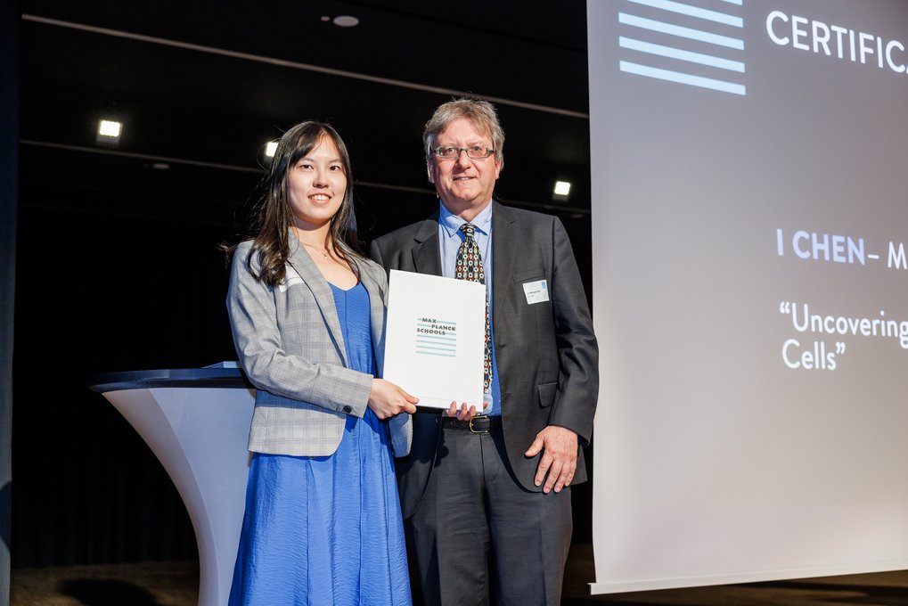 I Chen in a blue dress and gray blazer stands next to Jan-Michael Rost in a suit; I Chen holding a certificate on stage.