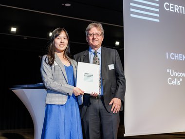 I Chen in a blue dress and gray blazer stands next to Jan-Michael Rost in a suit; I Chen holding a certificate on stage.