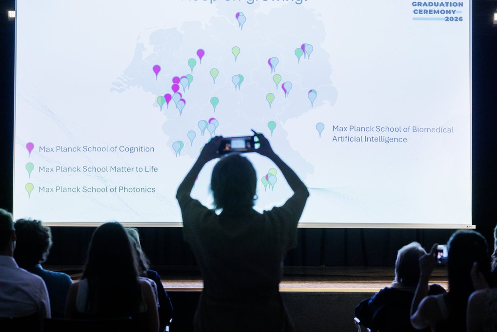 Attendees capture images of a projection displaying a map with colored markers indicating different Max Planck Schools across Germany during a graduation ceremony.