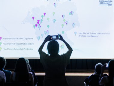 Attendees capture images of a projection displaying a map with colored markers indicating different Max Planck Schools across Germany during a graduation ceremony.