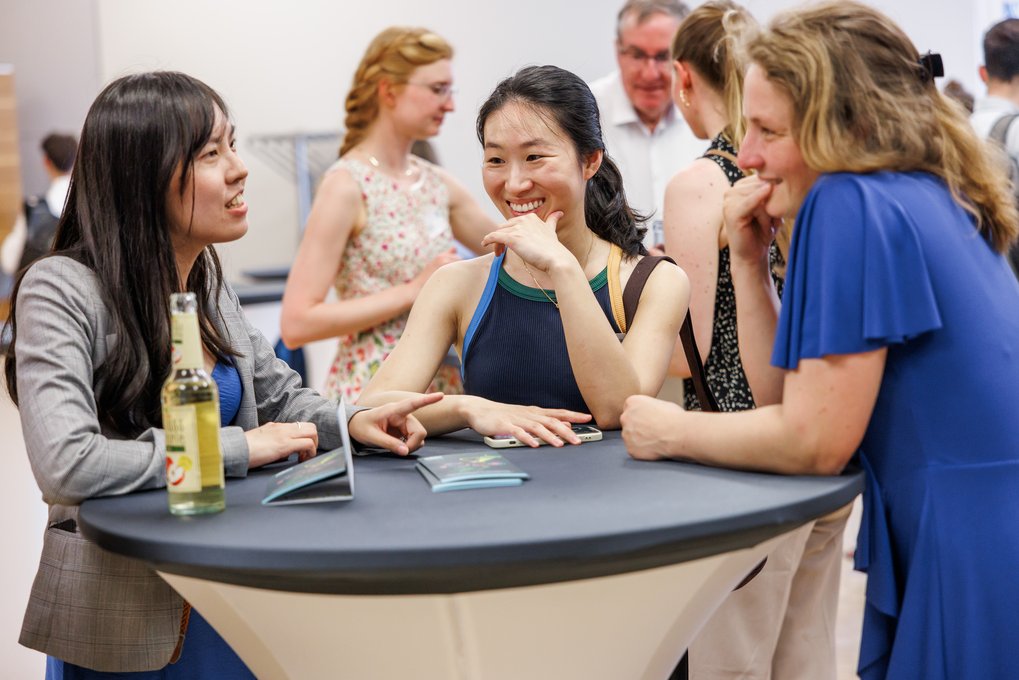 Three women engaged in conversation around a high table with drinks and brochures in a busy indoor setting.