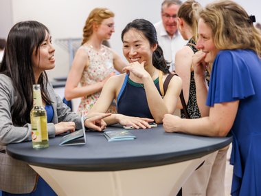 Three women engaged in conversation around a high table with drinks and brochures in a busy indoor setting.