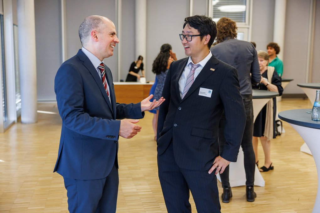 Two professionally dressed men engaged in conversation at a networking event with others in the background.
