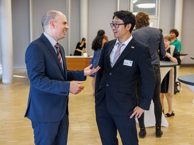 Two professionally dressed men engaged in conversation at a networking event with others in the background.