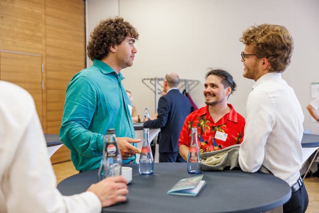A group of three men conversing at a high round table with bottled beverages, one wearing a turquoise shirt, another a red floral shirt, and the third in a white shirt with glasses.