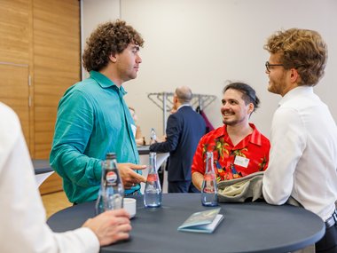 A group of three men conversing at a high round table with bottled beverages, one wearing a turquoise shirt, another a red floral shirt, and the third in a white shirt with glasses.