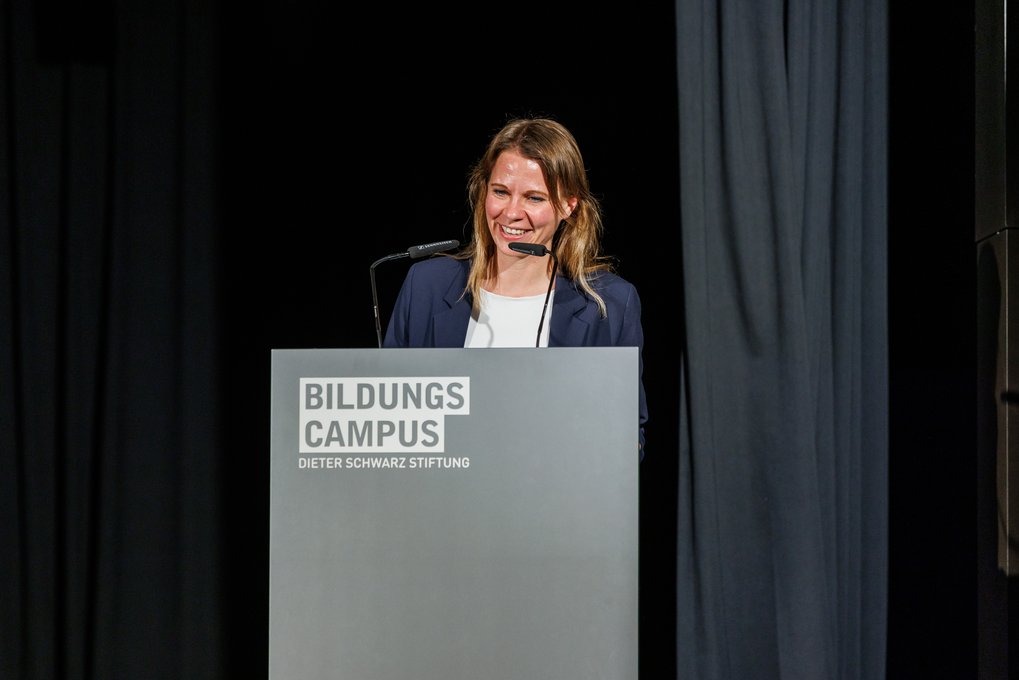 Miriam Biller in a dark blazer addresses an audience from a podium with the logo Bildungs Campus Dieter Schwarz Stiftung.