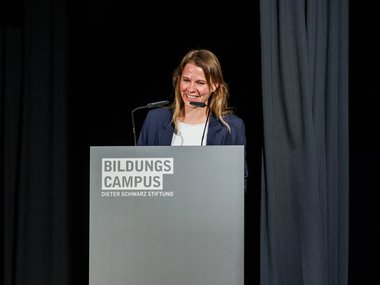 Miriam Biller in a dark blazer addresses an audience from a podium with the logo Bildungs Campus Dieter Schwarz Stiftung.