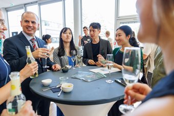 Doctors at last, officially celebrated: graduates raise a glass at the reception. Several Max Planck School graduates in business attire socialize and network at a standing table during the graduation ceremony, with drinks, snacks, and informational materials visible.