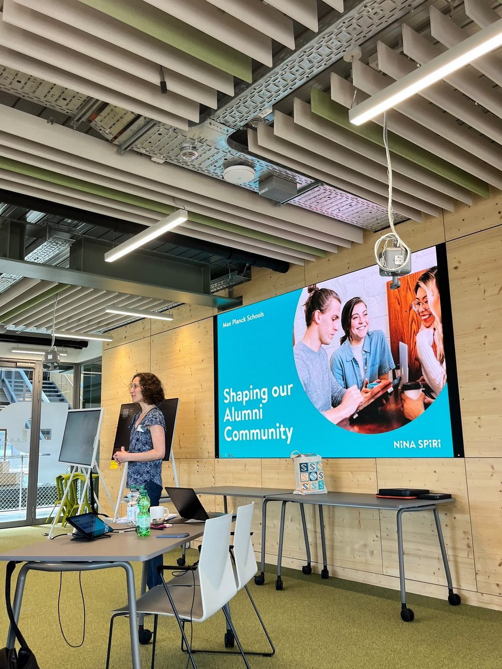 A woman giving a presentation in a conference room equipped with tables, chairs, laptops, and a large screen displaying "Shaping our Alumni Community" by Max Planck Schools.