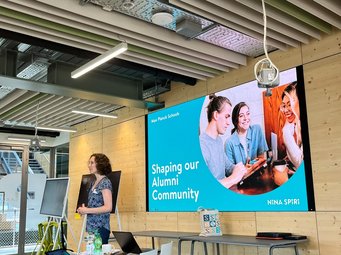 Setting the stage for the alumni discussion: moderator Nina Spiri kicks off the workshop. A woman giving a presentation in a conference room equipped with tables, chairs, laptops, and a large screen displaying "Shaping our Alumni Community" by Max Planck Schools.