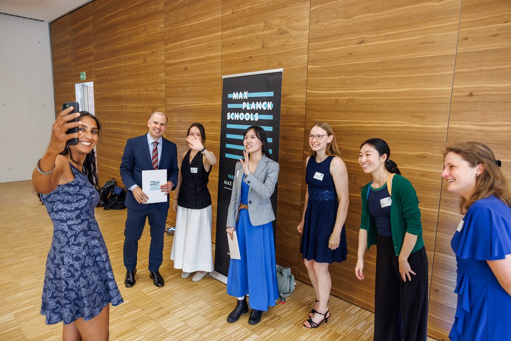 Diverse group of six people dressed formally and casually, gathered indoors by a Max Planck Schools banner, with one person taking a selfie and others smiling or waving.