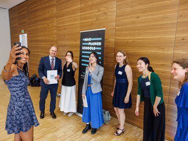 Diverse group of six people dressed formally and casually, gathered indoors by a Max Planck Schools banner, with one person taking a selfie and others smiling or waving.