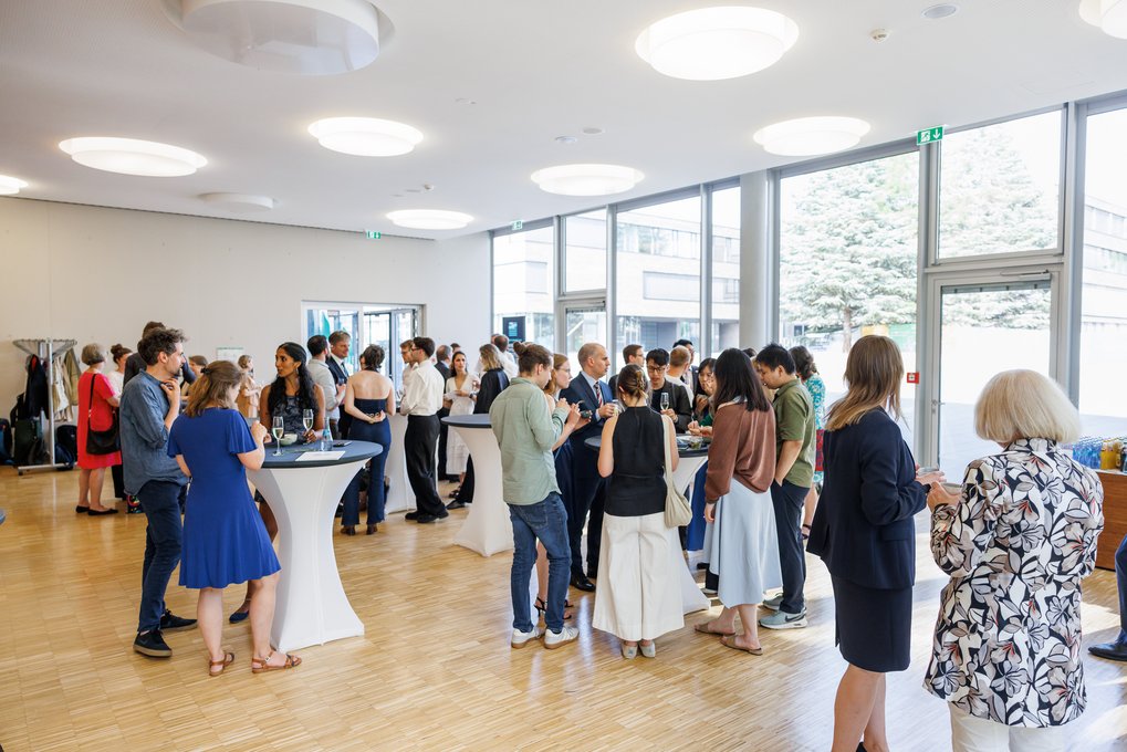 Numerous individuals gathered in small groups, talking and holding drinks at a standing reception inside a modern venue with wooden floors and large glass windows.