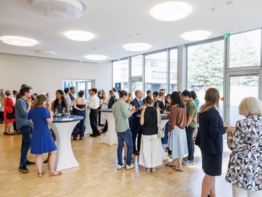 Numerous individuals gathered in small groups, talking and holding drinks at a standing reception inside a modern venue with wooden floors and large glass windows.