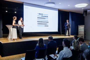 Two women and one man presenting on stage with an audience in front and a screen displaying the Max Planck School Logo with letters "Max Planck School Graduation Ceremony 2026"
