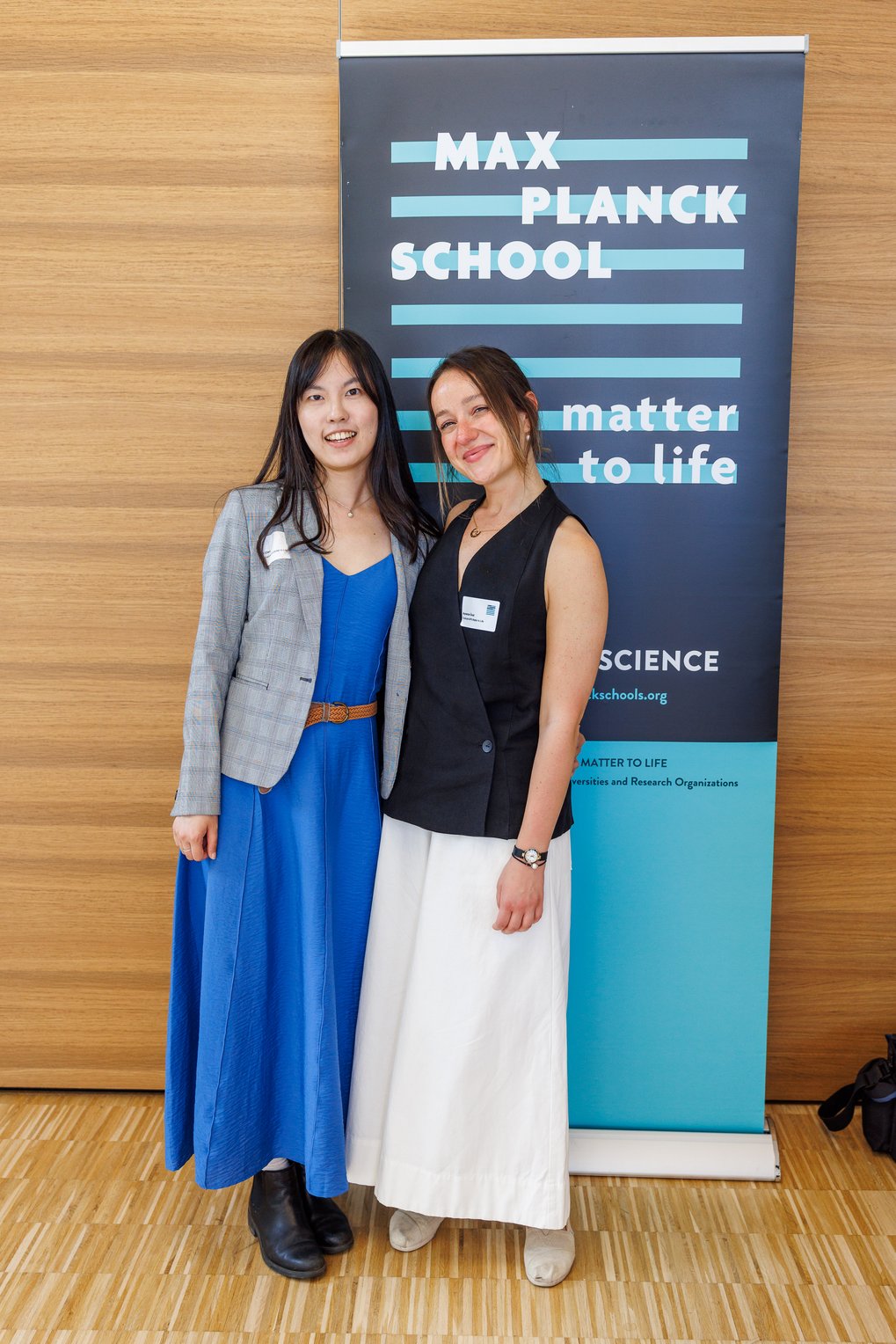 Aysecan Ünal and I Chen wearing formal attire standing side by side, smiling, with a Max Planck School banner behind them and a wooden wall background.