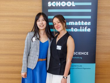 Aysecan Ünal and I Chen wearing formal attire standing side by side, smiling, with a Max Planck School banner behind them and a wooden wall background.