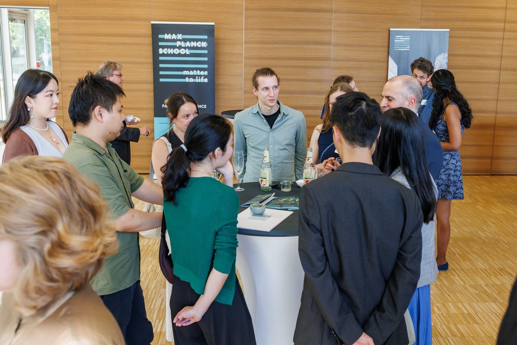 Attendees gathered around a cocktail table during a social event, with beverages and informational materials visible, set against a wooden-paneled background.