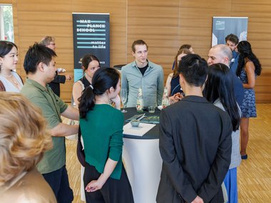 Attendees gathered around a cocktail table during a social event, with beverages and informational materials visible, set against a wooden-paneled background.