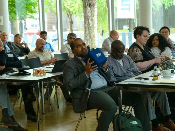 A diverse group of people attending a conference or workshop in a room; some are taking notes or listening, while a man is clearly seen holding a blue cube-shaped microphone and speaking into it.
