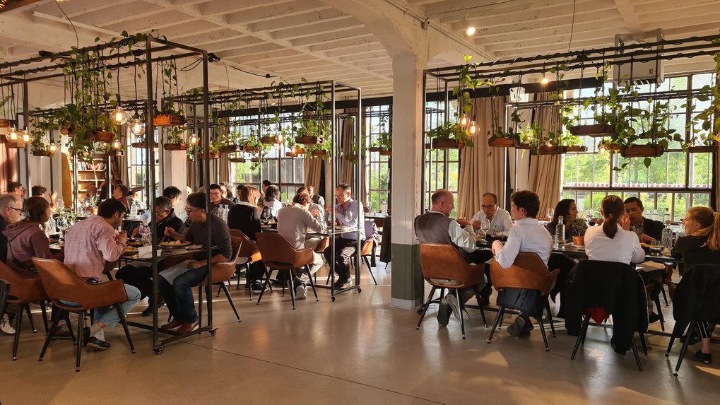 Several diners in a modern, well-lit restaurant with large windows and suspended plant arrangements above the tables.