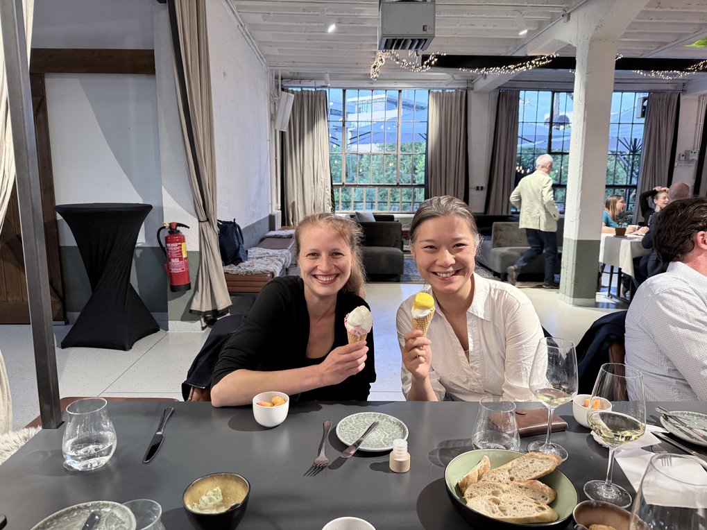 Two women enjoy ice cream cones while seated at a dining table with plates, glasses, and bread in a spacious, modern room with large windows.