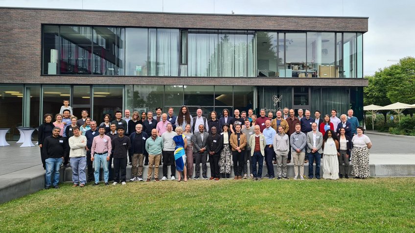 Fifty researchers, two continents, three days in Heilbronn – the second joint Matter to Life Conference with the Sloan Foundation, June 9–11, 2026. Diverse group of about 50 people posing for a photograph on a grassy area outside a modern office building featuring extensive glass facades and brickwork.