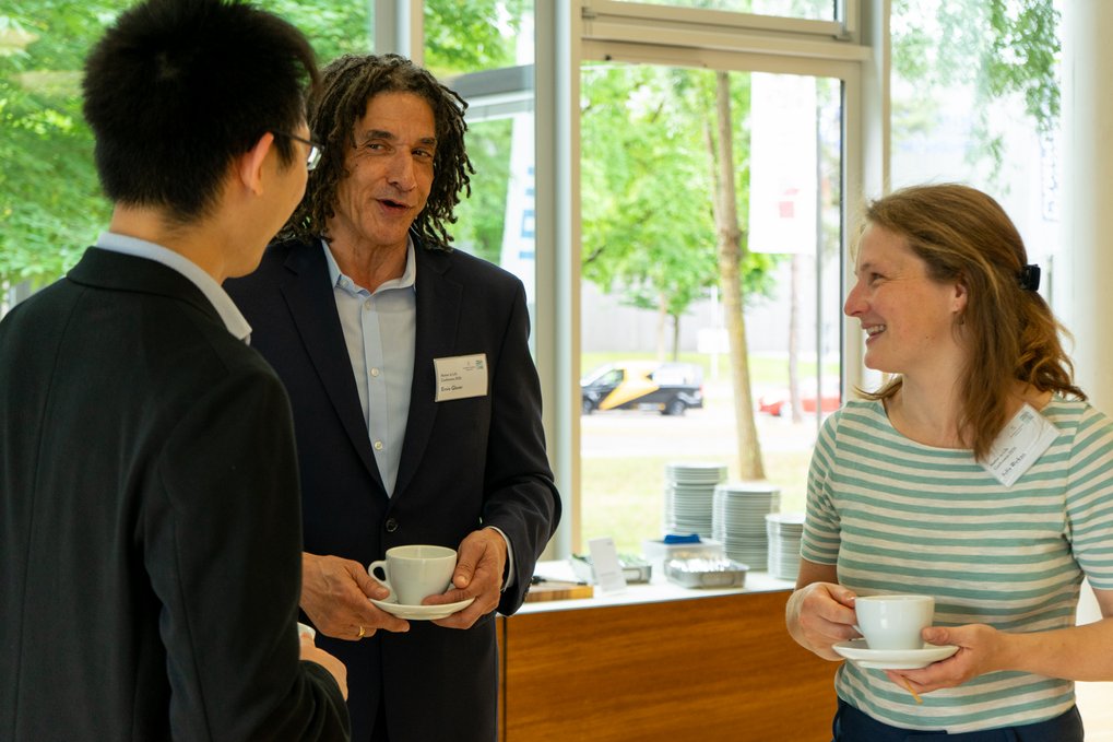 A group of three people, two men and one woman, talking and holding coffee cups in a bright room with large windows and a table with plates behind them.