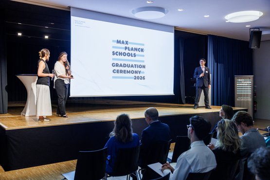 At the Max Planck Schools Graduation Ceremony 2026, three of the graduates address an audience from a stage, with a large screen displaying the event name behind them.