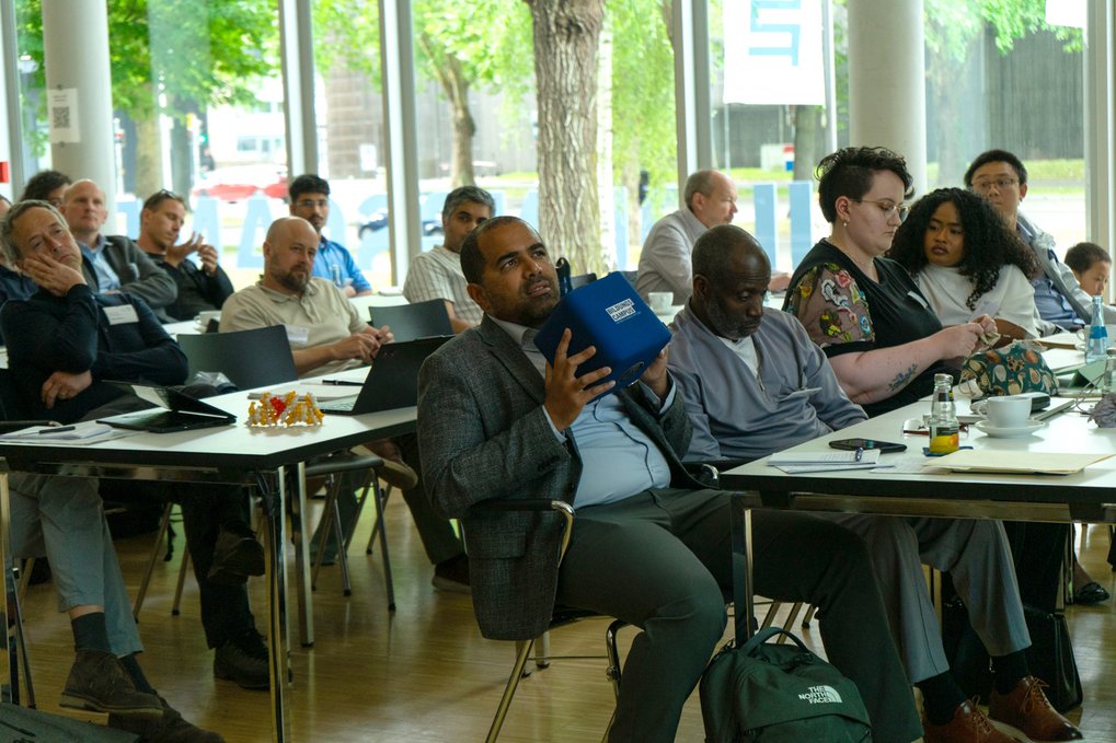 A diverse group of adults is listening intently to a lecture in a bright seminar room with large windows and modern amenities.