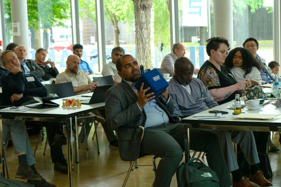 A diverse group of adults is listening intently to a lecture in a bright seminar room with large windows and modern amenities.