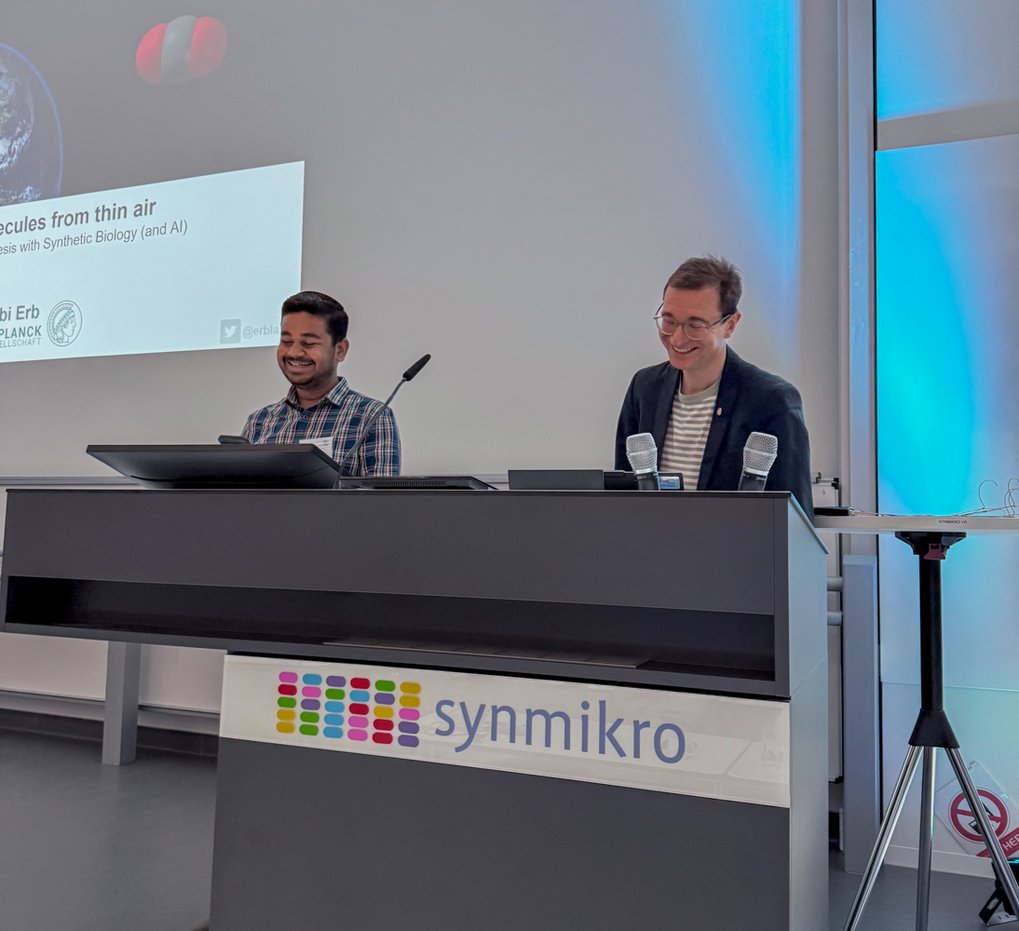 Two men standing behind a lectern with the sign "synmikro" in a conference room.