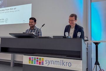 Two men standing behind a lectern with the sign "synmikro" in a conference room.
