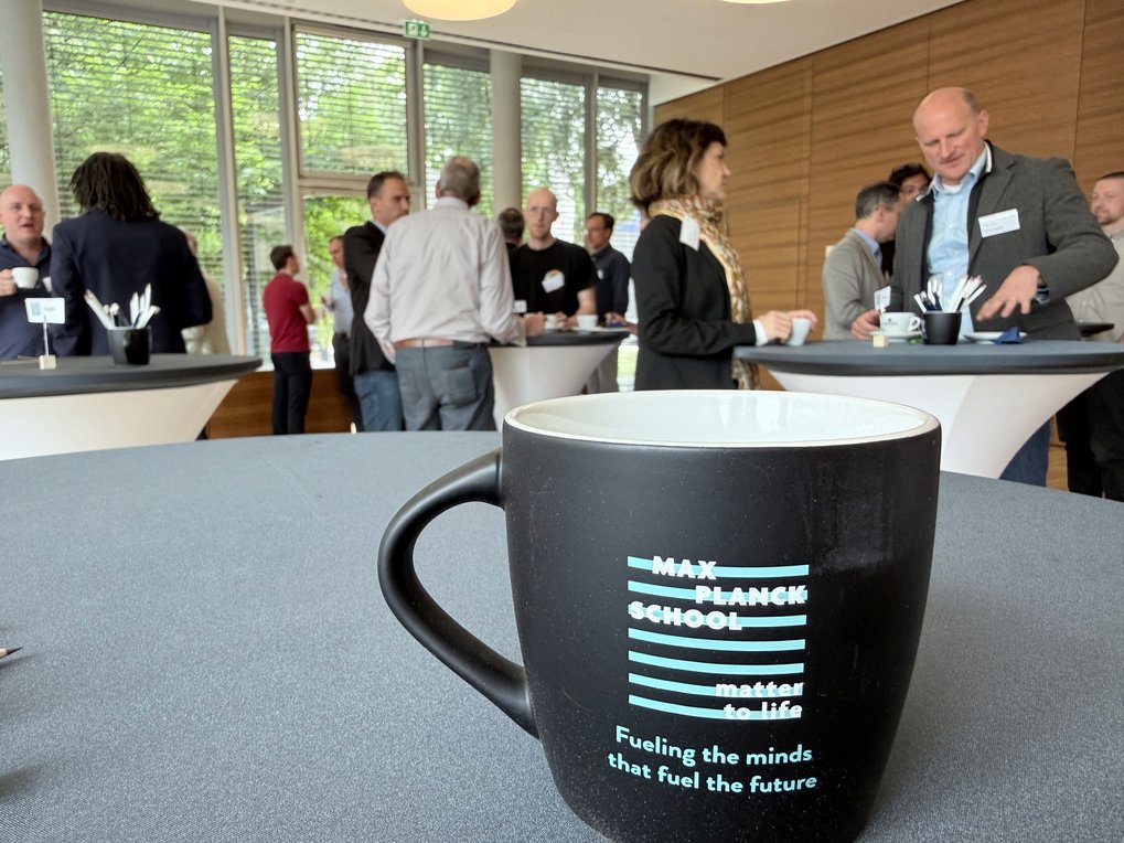 Coffee mug branded with Max Planck School Matter to Life logo on a table in the foreground, several people networking near high tables in a bright room.
