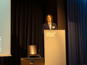 Ernie Glover welcoming participants from both sides of the Atlantic to the second joint Matter to Life Conference Ernie Glover standing at a podium on a stage with a presentation screen to the side.