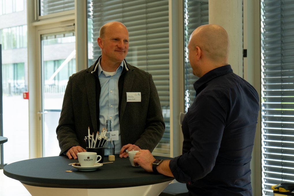 Two men in business casual attire conversing at a round table with coffee cups, pens, and a water bottle, in a brightly lit room with large windows and blinds.
