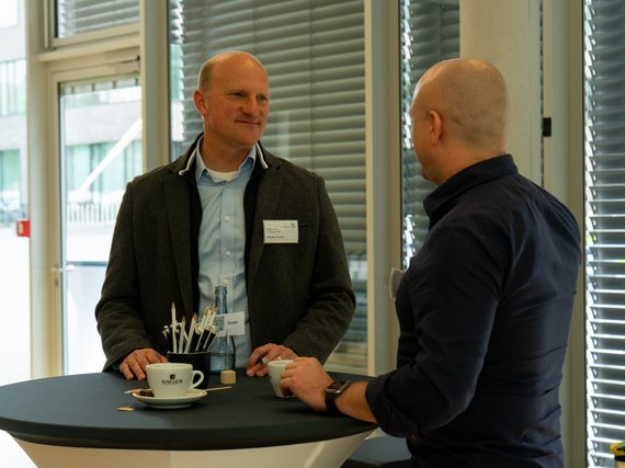 Two men in business casual attire conversing at a round table with coffee cups, pens, and a water bottle, in a brightly lit room with large windows and blinds.