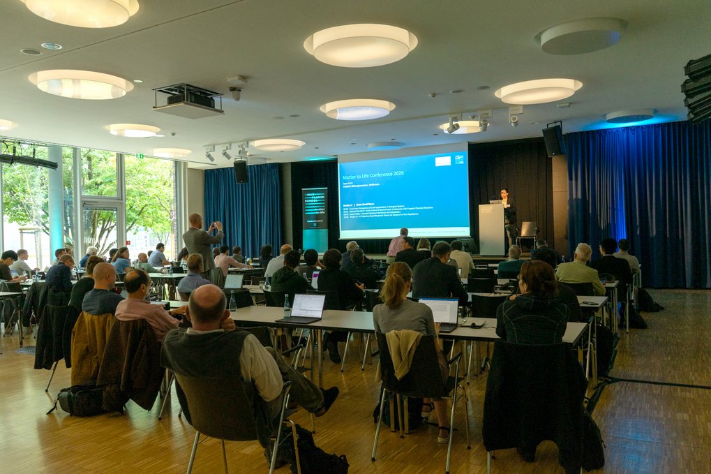Audience seated at tables in a conference room listening to a speaker presenting data visuals on a screen.