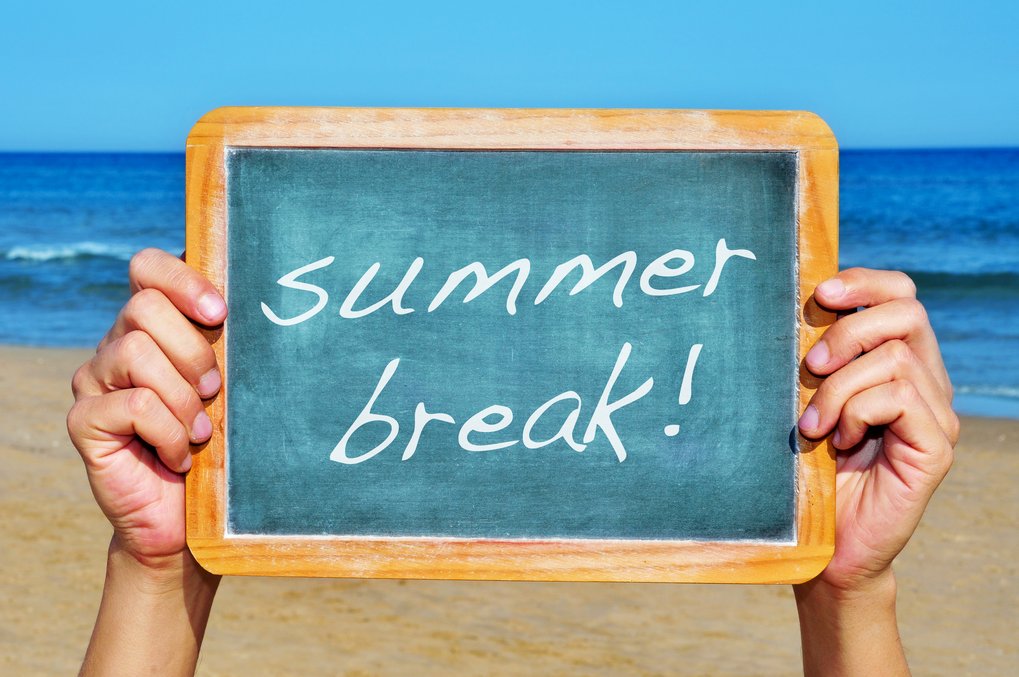 Small chalkboard with the text "summer break!" held by hands in front of a sandy beach and ocean background.