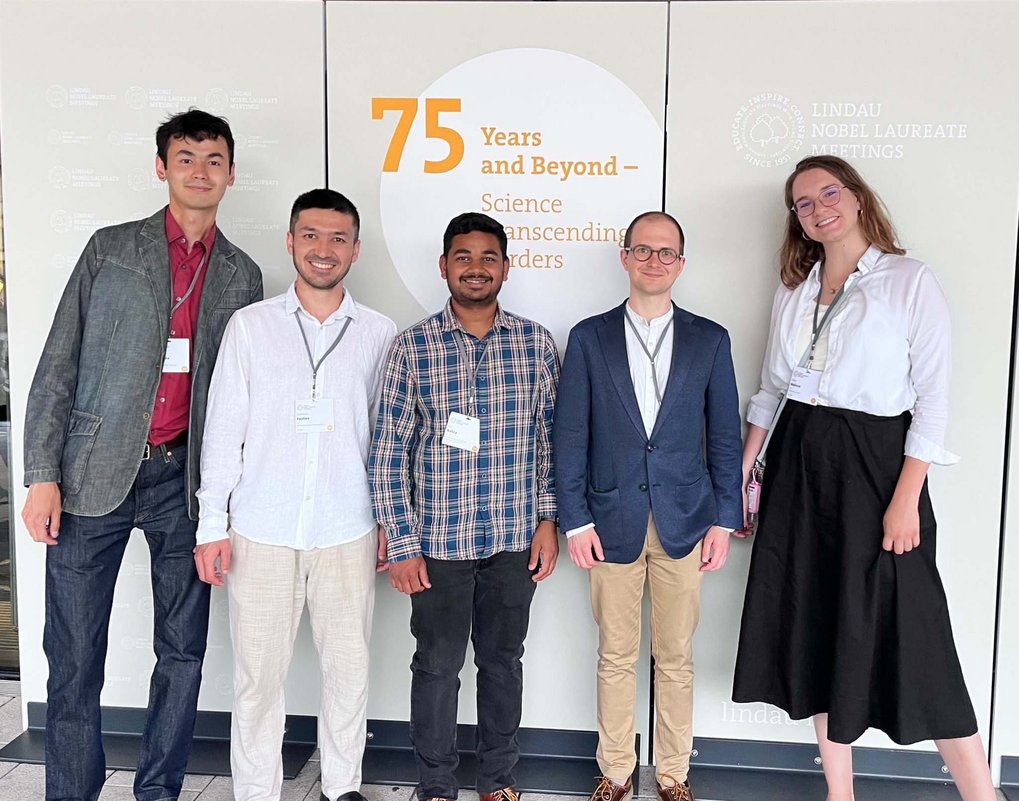A group of five MtL Ph.D. candidates posing in front of a wall displaying the words “75 Years and Beyond – Science Transcending Borders” and the logos of the Lindau Nobel Laureate Meetings.