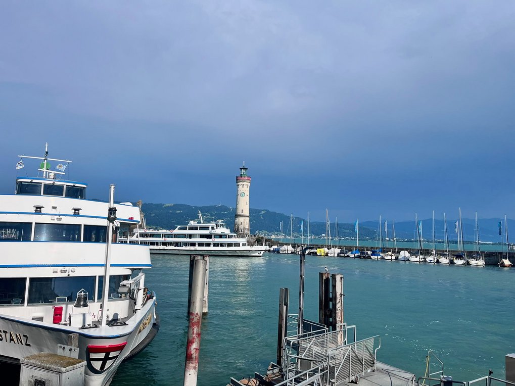View of a harbor featuring several boats docked near a lighthouse with mountains in the background.