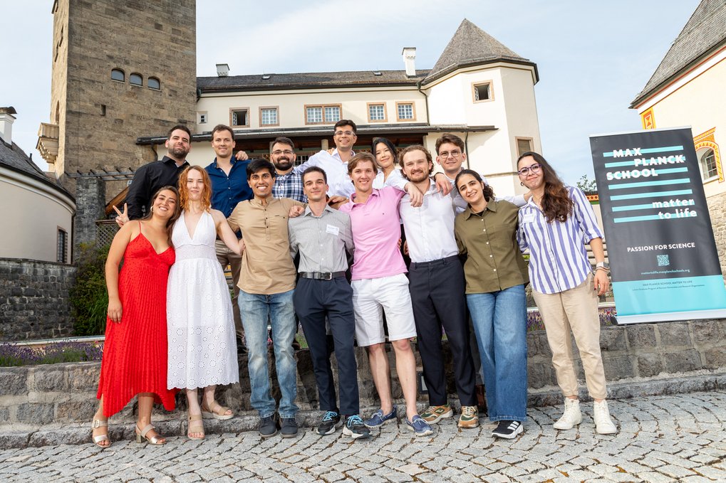 Thirteen Matter to Life PhD candidates in casual attire standing on cobblestone steps, smiling and linking arms in front of a historic castle-like structure and a sign reading "Max Planck School – Passion for Science."