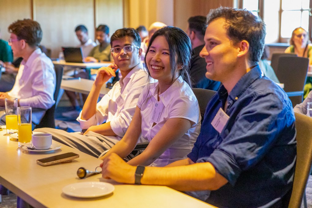 Two participants wearing badges at a table during an event, with other attendees in the background.