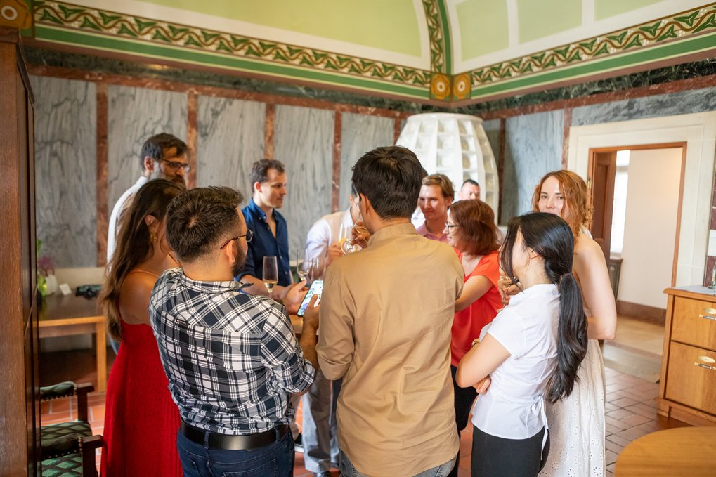 A diverse group of people standing in a decorated room, interacting socially with drinks in hand, some looking at a phone.