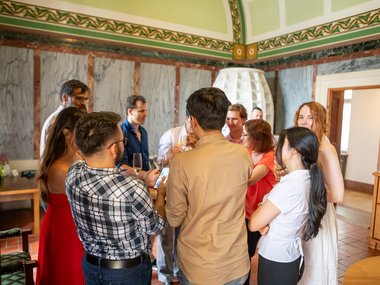 A diverse group of people standing in a decorated room, interacting socially with drinks in hand, some looking at a phone.