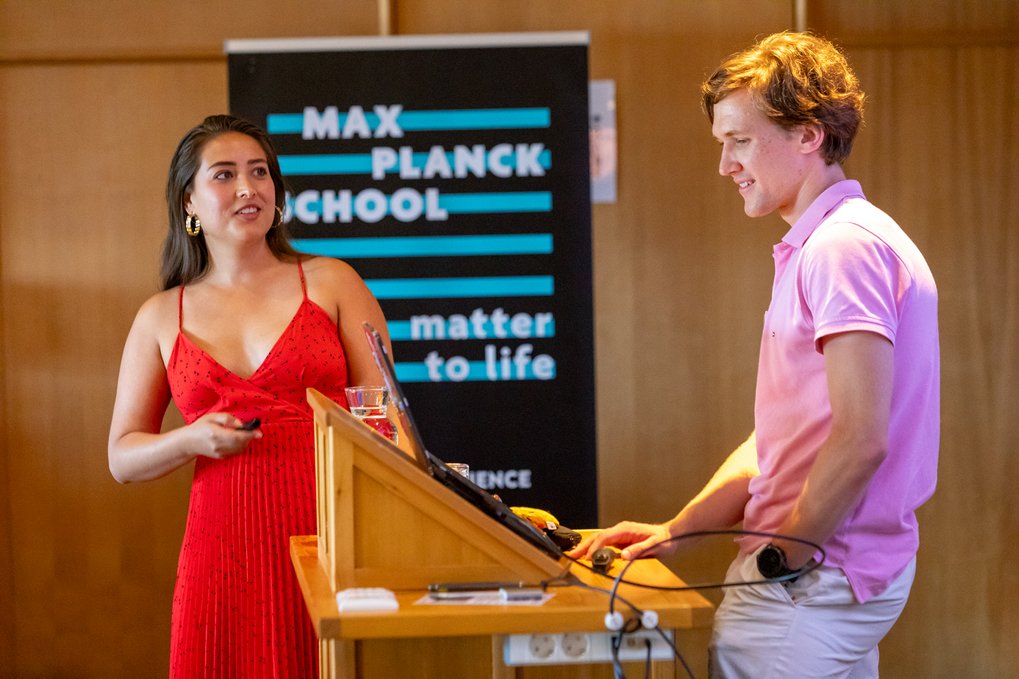 A female presenter in a red dress speaks while a male colleague operates a laptop at a lectern, with a Max Planck School Matter to Life banner in the background.