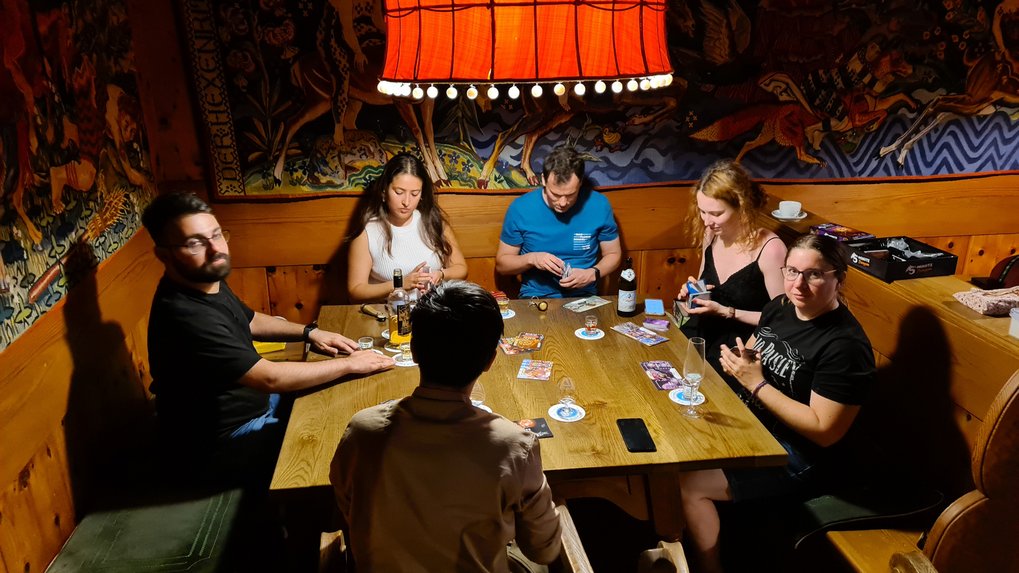 Six individuals gathered around a wooden table in a cozy room, focused on playing a card game with drinks and game materials visible.
